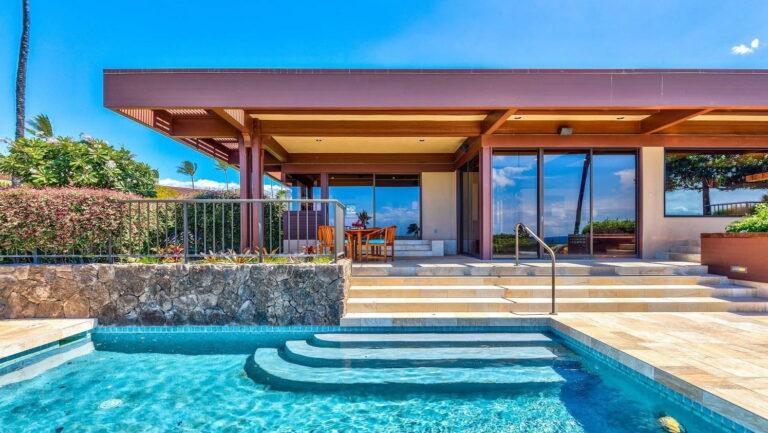 Modern tropical home beside a turquoise pool, with curved steps, glass doors, shaded patio, and lush greenery creating a serene feel under blue skies.